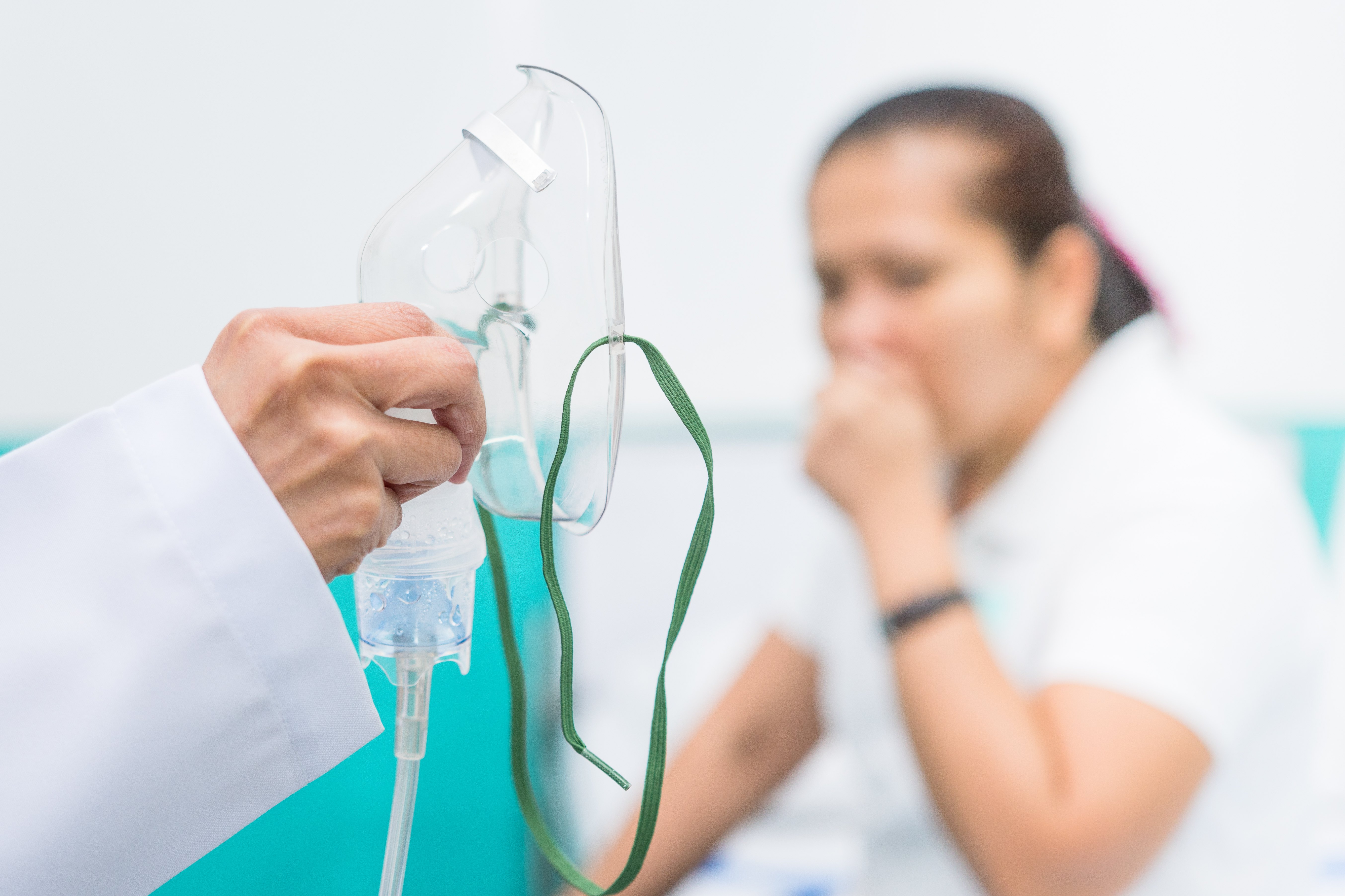 Respiratory therapist holds an oxygen mask with a nebulizer as a coughing patient watches in the background.