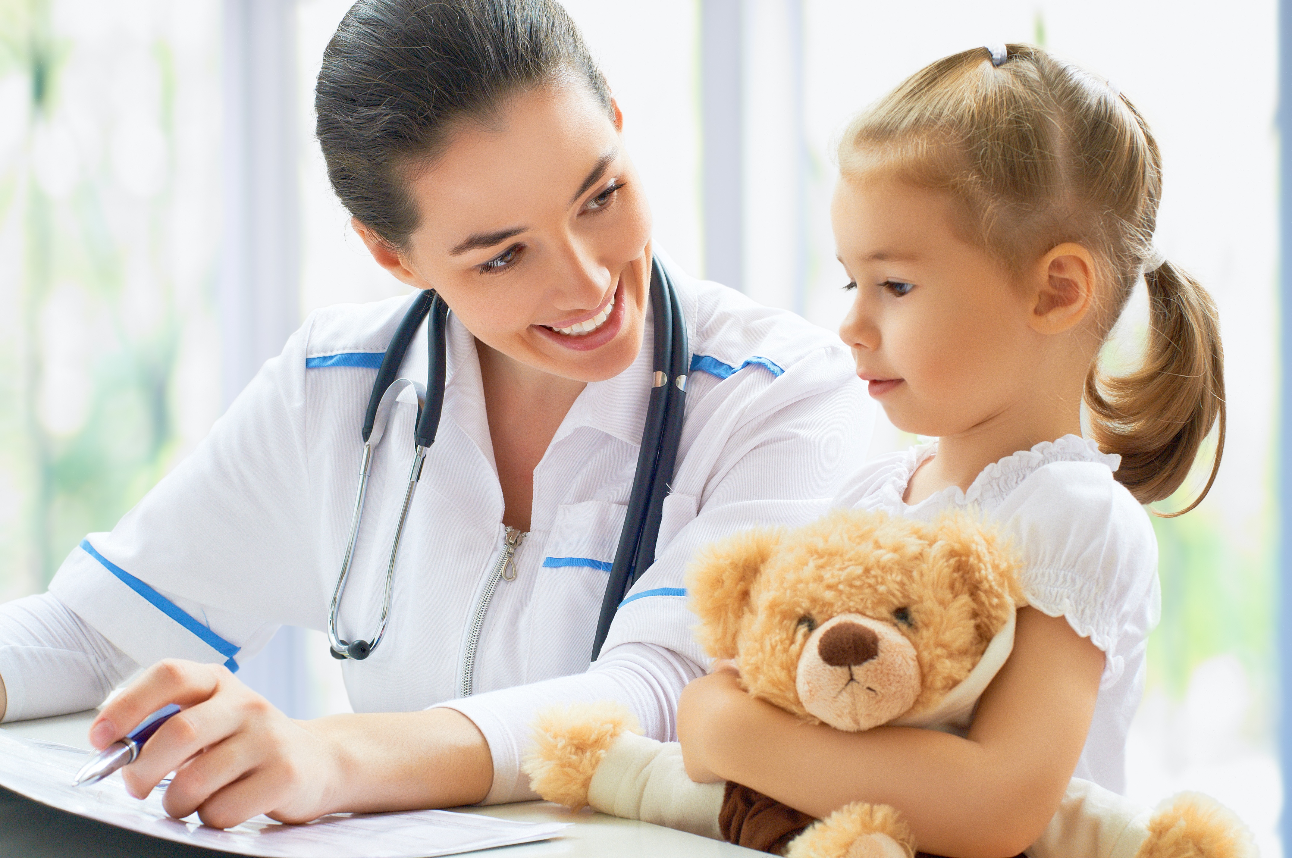 physician smiles at her young patient who is holding a teddy bear